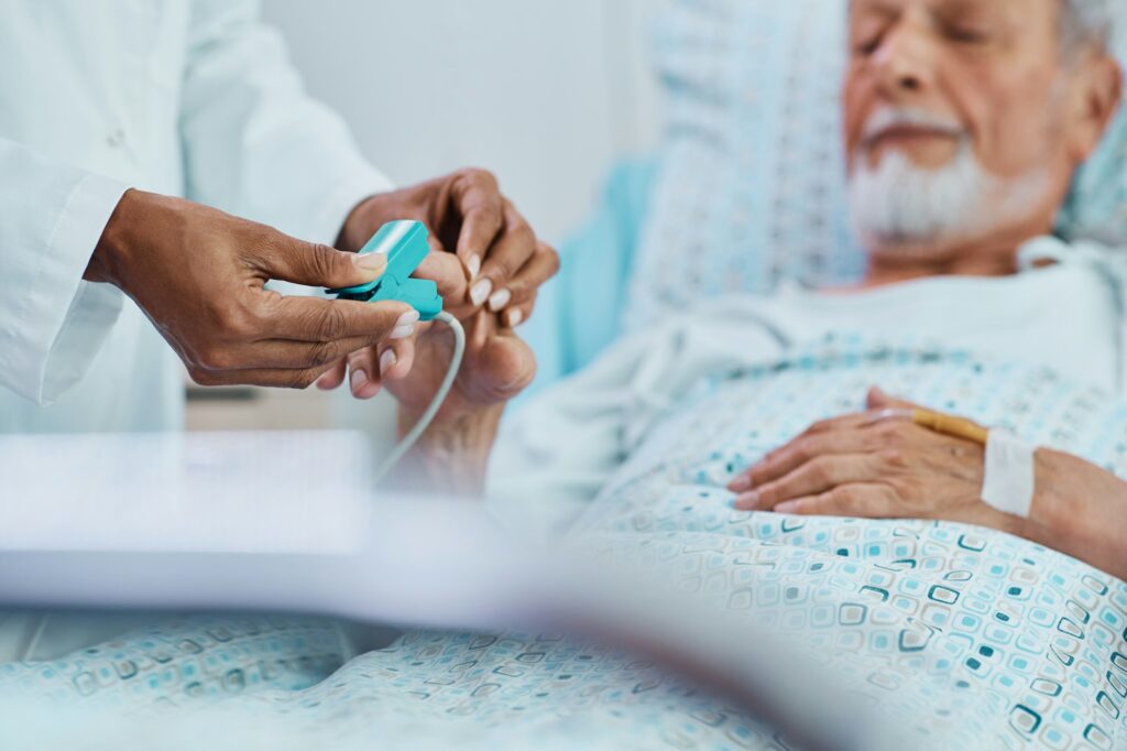 Close up of black doctor putting pulse oximeter on finger of senior man in hospital recovery unit.